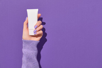 Womans hand holding white tube on violet background. Skin health care concept.
