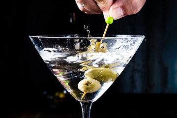 Hand removing some olives from a classic conical martini glass and generating splashes. Horizontal orientation.