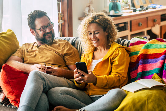 Adult couple at home enjoy mobile phone activity sitting on the sofa and smiling. Colorful house interior and cheerful man and woman people having morning breakfast with technology