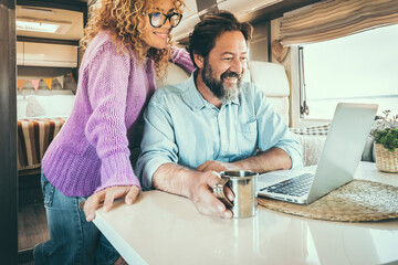 Adult couple use laptop computer sitting inside a camper van and smile. Nomadic online lifestyle people enjoy internet connection inside a motorhome camping car with beach outside the window - obrazy, fototapety, plakaty