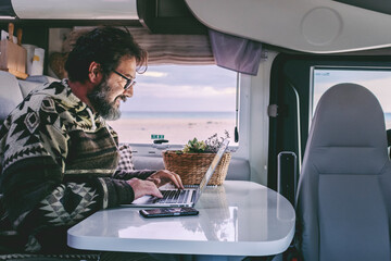 Adult man use laptop computer to work inside camper van with roaming phone connection. Concept of...