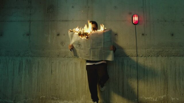 Footage of male artist with sunglasses standing near wall , holding and reading newspaper during it burns . Concept or symbol of fake or forbidden news   . Man reads burning magazine . Slow motion
