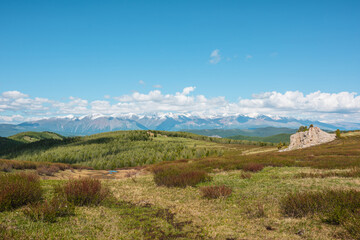 Atmospheric landscape with big rock on hill with view to sunlit mountain vastness and high snowy mountain range under low clouds line on horizon. Impressive scenery with vast mountains in sunlight.