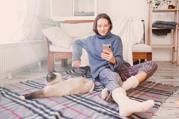 Girl sitting near sofa holding phone in her hands in an warm sweater on the floor