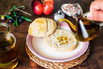 Hand of a man with an oil can pouring extra virgin olive oil into a bread roll next to a tomato and garlic.