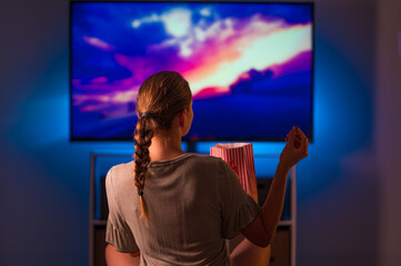 A fair-haired girl in home clothes watches TV with a pack of popcorn. Blue neon light in the room....