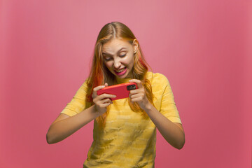 Gambling young stylish modern girl playing online game using mobile phone app, grimacing on pink studio background