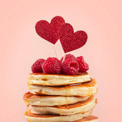 Close-up of stack of fluffy pancakes with syrop, decorated with red glitter papper hearts and srtawberries. Square format. Light peach colour background. Copy space.