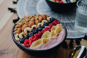 bird’s eye view of a vegan berry smoothie bowl with fruits and granola like crunshy oats, raspberries, blueberries, bananas, daisies, cashew nuts on a wooden table for a healthy breakfast with a spoon