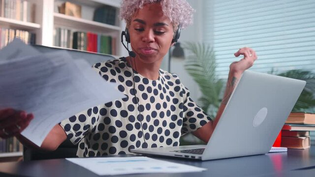 Attractive successful African American woman with call center headset on head works in psychological support service or social consultation through laptop and ip telephony sits at office desk