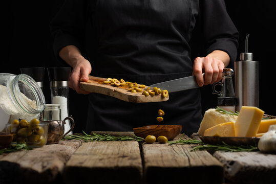 A chef in a black uniform cuts green olives on a cutting board. Ingredients for making salad, soup, sauce, focaccia, pie. Wooden texture, dark background. Restaurant, hotel, recipe book.