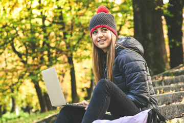 Girl with wool cap sitting in the park using laptop, beautiful autumn afternoon. Teen use computer to communicate with friends and classmates. Young student woman enjoying free time in nature