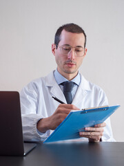 Young doctor man with eyeglasses writes on clipboard for his work, doctor sitting in front of his desk with laptop