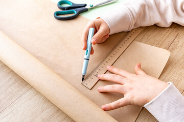 Close-up of child's hand in process of sketching with pencil on craft paper. Straight line with ruler. Crafts and packaging from eco paper. School Child or preschooler doing homework. Hobby, origami