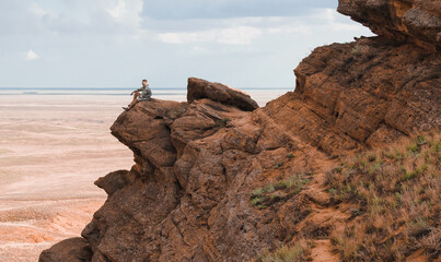 Man from the side, sitting on top of a rock, looking to distance beyond the horizon.Red mountain...