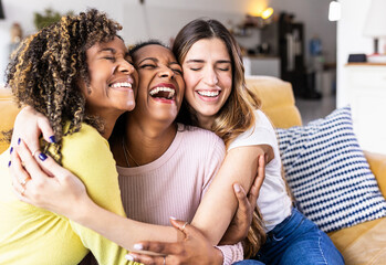 Three happy multiethnic female best friends laughing together - Diverse young women hugging each other and having fun at home - Unity concept - Powered by Adobe