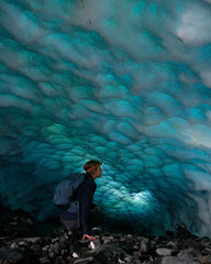 Adventurous Ice Climber Woman walking in an ice cave on a glacier in Alaska. Glacier is receding and melting due to global warming. Dark blue ice and headlamp - obrazy, fototapety, plakaty