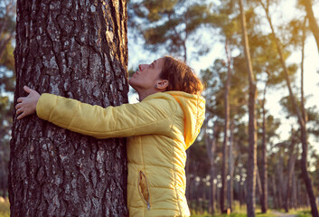 young girl with yellow jacket in profile hugging a tree in a pine forest at sunset