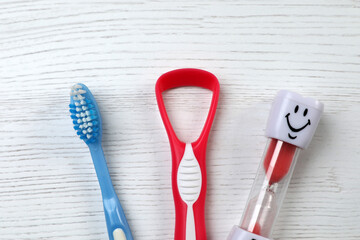 Tongue cleaner, toothbrush and hourglass on white wooden table, flat lay