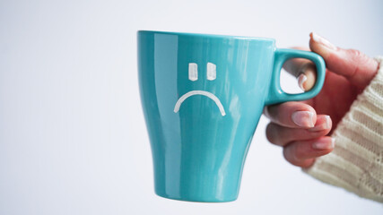 Blue Monday concept. Close-up of a female hand in a white sweater holds a blue cup with a sad smiley as a symbol of the most depressive day of the year on a white background.
