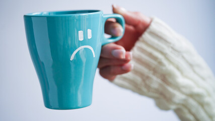 Blue Monday concept. Close-up of a female hand in a white sweater holds a blue cup with a sad smiley as a symbol of the most depressive day of the year on a white background.