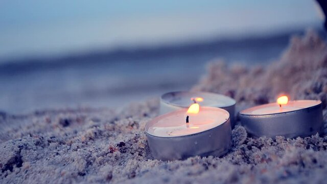Three small burning candles on sand beach on background of blurry sea waves close-up. Candlelight, candle flame. Romantic mood backdrop. Concept date relaxation leisure vacation. Sandy shore sea coast