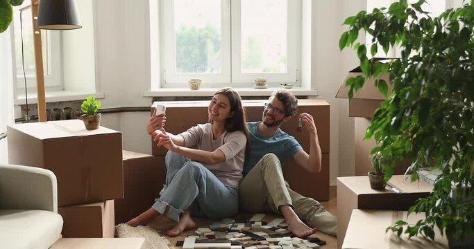 Happy couple sit on floor near cardboard boxes with personal stuff, use smart phone take selfie, show keys, make picture to remember day of moving to own house. Tenancy, modern tech, relocate concept