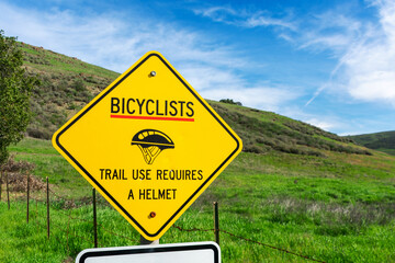 Bicyclists trail use requires a helmet yellow warning sign. Blurred green hills background under...