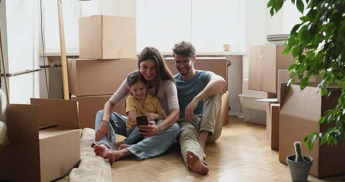 Couple homeowner family their little daughter using cellphone sit on floor near carton boxes with stuff on relocation day, resting use mobile app, have fun on-line. Modern tech usage, new home concept