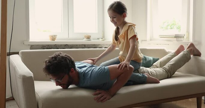 Little 5s girl sits rides on daddy back play together at home. Young loving father lying on cozy couch in living room enjoy playtime with adorable preschool daughter. Family bond, funny games concept