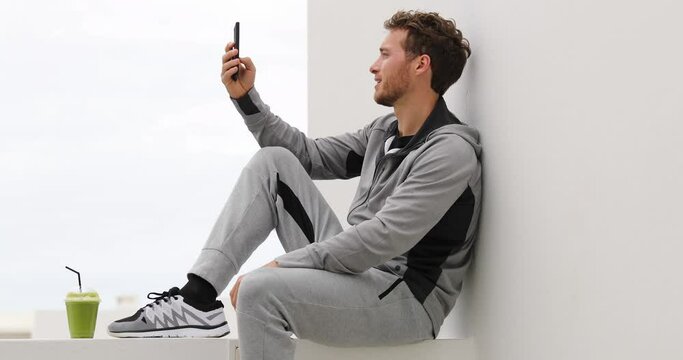 Phone. Young man using smartphone watching online video or texting sms messages. Fitness man using phone app on running break relaxing outdoors. Green smoothie and sweatpants healthy active lifestyle