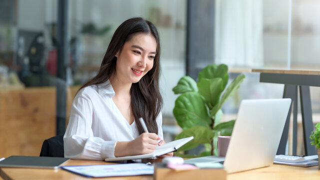 Confident Asian businesswoman sitting and taking notes with a laptop computer in the office happily.
