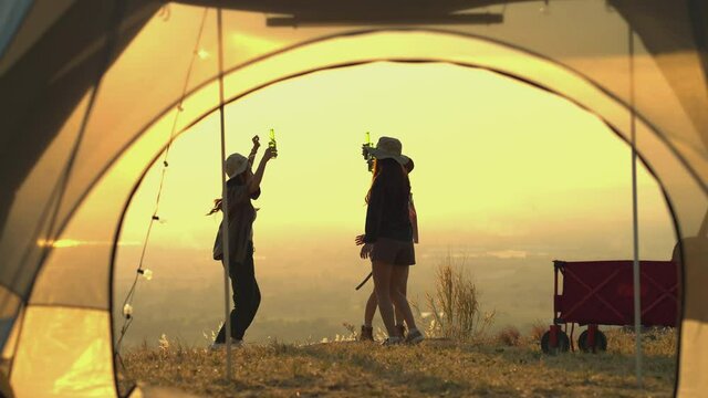 Friendship group of Asian young women having fun and enjoy partying. They are all holding bottles of beer and celebrating on the mountain outdoor camping trip in nature during sunset. slow motion
