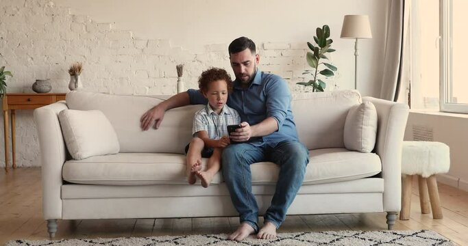Dad and 5s son sit on sofa with smart phone spend free time together use modern wireless tech and internet connection, father teach kid use technology. Gadget overuse, bad habit, new app usage concept