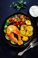 Fried salmon steak with potatoes and fresh vegetable salad served on black plate on wooden table
