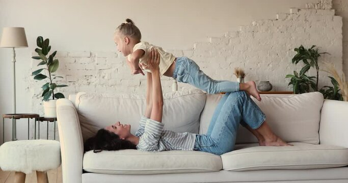 Cheerful woman lifts on arms her cute little daughter while lying on sofa, play with preschool 4s kid, do acroyoga together at home. Playtime with children, happy motherhood, custody, foster concept
