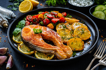 Fried salmon steak with potatoes and fresh vegetable salad served on black plate on wooden table

