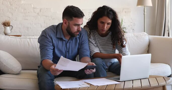 Couple sit on couch at table with laptop pay bills, use calculator count expenses, make budget control, manage finances, review overdue loan payments feel stressed. Crisis, lack of money, debt concept