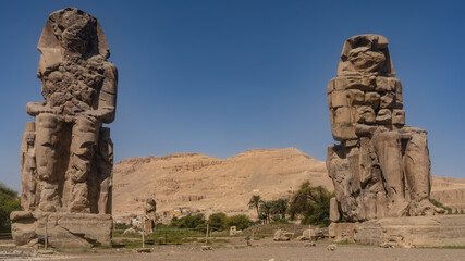 Giant sculptures of the colossi of Memnon against a blue sky and a sand dune. Huge statues of seated pharaohs are dilapidated. Egypt