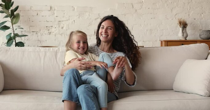 Hispanic woman her cute little daughter have fun smile look at camera, enjoy playtime sit on sofa at home. Babysitter tickling preschool adorable girl play together in living room. Family bond concept