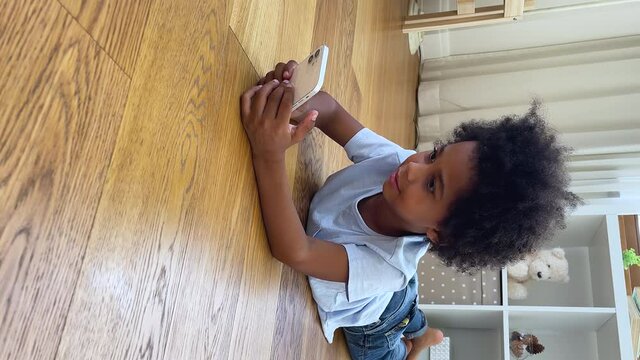 Vertical view adorable junior school age African American boy lie on wooden floor alone play internet game on smartphone. Little child addicted to gadget surf social media watch popular vlogs online