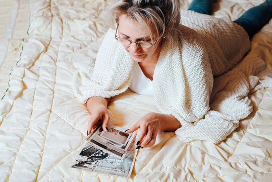 Nostalgia and memories concept. Middle-aged caucasian woman looking at old family album photos while relaxing on bed in bedroom, top view. Adult woman and vintage pictures of childhood and youth