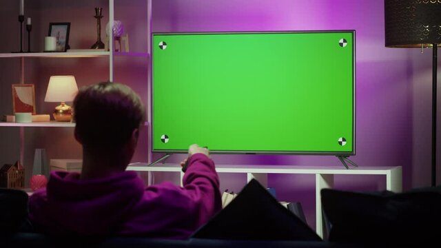 Man watching television in living room in evening. Young guy sitting on sofa against tv with chroma green screen, back view. Male person switching channels using remote controller.