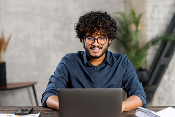 Front view of positive Hindi man in smart casual shirt using laptop while sitting at the desk in his flat. Young Indian male student watching webinars, educational courses, learning on the distance
