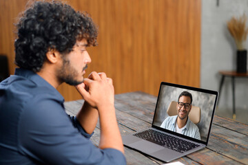 Back view of the Indian guy talking with male friend or coworker via video call. He sitting at the office desk and using laptop for video meeting. Young man on the screen