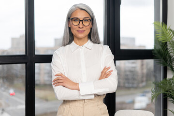 Serious and strict old senior business woman wearing smart casual shirt and stylish eyeglasses standing with arms crossed in modern office space. Mature bossy lady, elderly purposeful leader concept