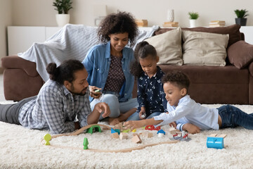 Happy Black couple of parents helping two kids to build toy road model, playing with wooden...