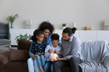 Happy millennial African American parents teaching little sibling kids to save money, making reserve. Family couple with son and daughter dropping cash, putting money into piggybank - Powered by Adobe