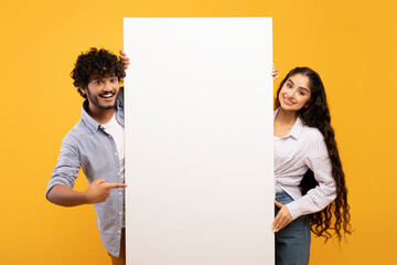 Happy indian couple in love standing by white empty board for advertisement or text over yellow...