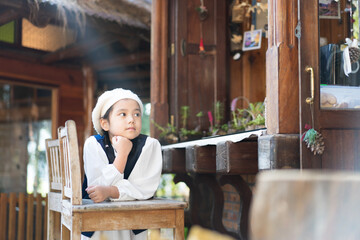 asian child tourist or kid girl cute wearing hat and vintage fashion dress or outfits to take a portrait photo with wooden chair and sunbeam on morning in coffee shop or restaurant on holiday travel